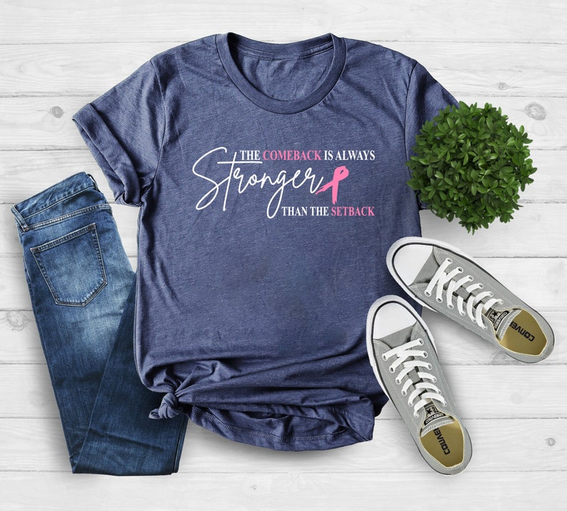May include: A navy blue t-shirt with the text "The Comeback Is Always Stronger Than The Setback" in white and pink. The shirt is paired with blue jeans and gray sneakers.