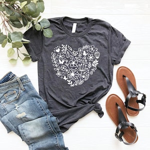 May include: A dark gray t-shirt with a white floral heart design. The heart is made up of flowers, leaves, and butterflies. The shirt is laid flat on a white wooden surface.