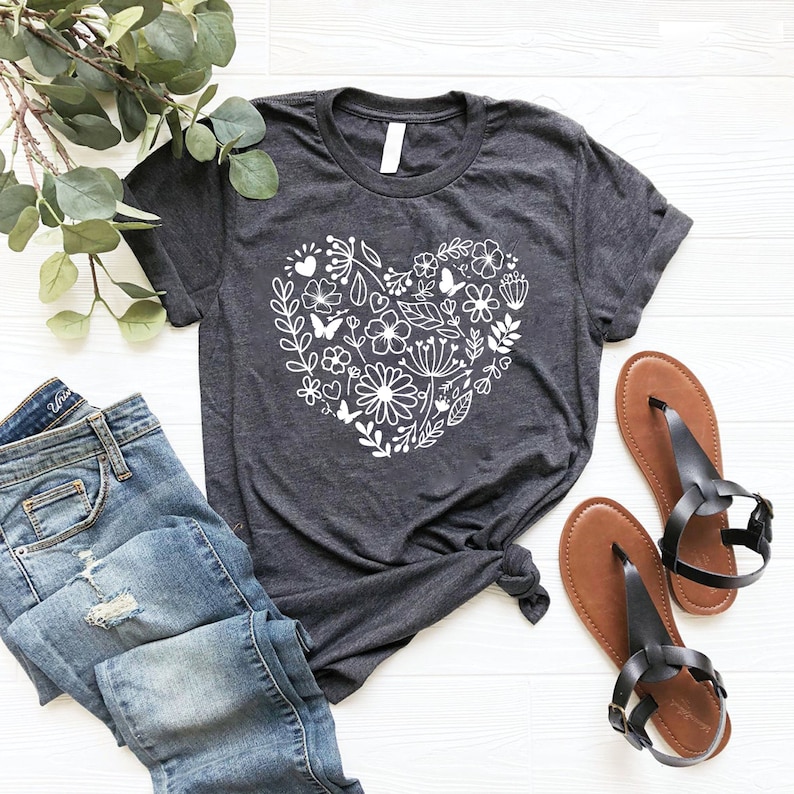 May include: A dark gray t-shirt with a white floral heart design. The heart is made up of flowers, leaves, and butterflies. The shirt is laid flat on a white wooden surface.