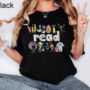 May include: A black t-shirt featuring the word read in white text, surrounded by colorful illustrations of various children's book characters including crayons, a cat, a monkey, an elephant, a pig, and other animated figures.