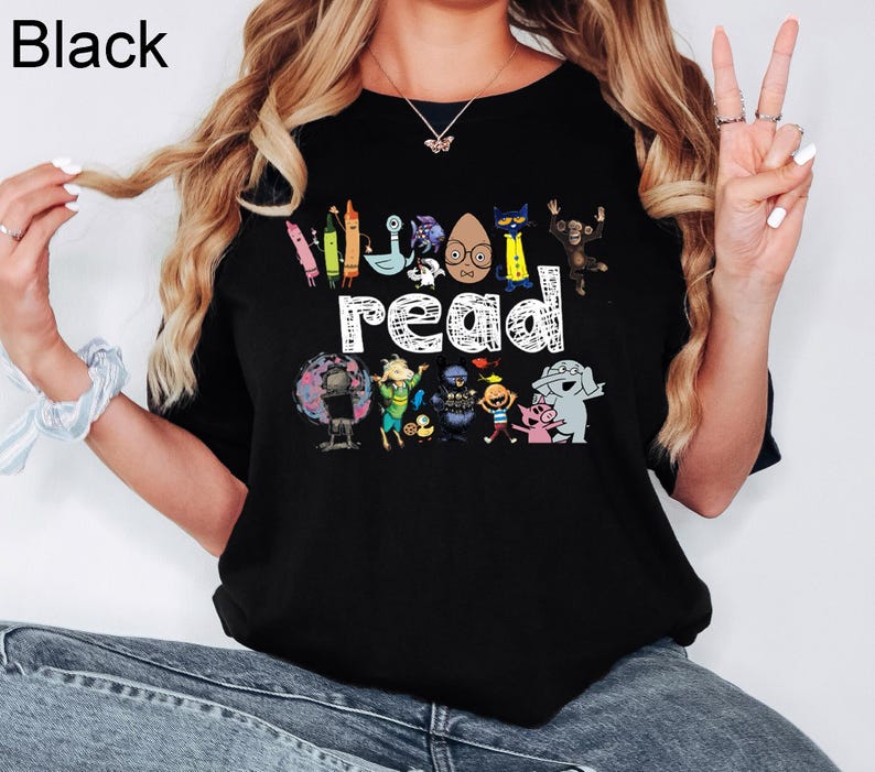 May include: A black t-shirt featuring the word read in white text, surrounded by colorful illustrations of various children's book characters including crayons, a cat, a monkey, an elephant, a pig, and other animated figures.