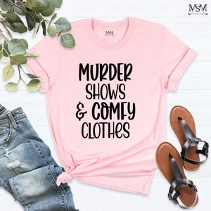 May include: A light pink t-shirt with the text "MURDER SHOWS & COMFY CLOTHES" in black. The shirt is paired with blue jeans and brown sandals with black straps. The t-shirt is a casual wear item.