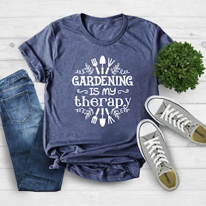 Op de afbeelding: Een marineblauw t-shirt met witte tekst die "Gardening is my therapy" zegt en een ontwerp van tuingereedschap en bladeren. Het shirt ligt op een wit houten oppervlak met een paar grijze Converse-schoenen en een kleine groene plant.