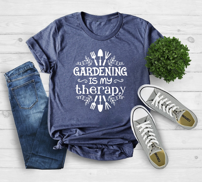 Op de afbeelding: Een marineblauw t-shirt met witte tekst die "Gardening is my therapy" zegt en een ontwerp van tuingereedschap en bladeren. Het shirt ligt op een wit houten oppervlak met een paar grijze Converse-schoenen en een kleine groene plant.