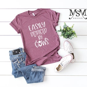 May include: A maroon t-shirt with the text "EASILY DISTRACTED BY COWS" in white. The shirt is paired with blue jeans and white sneakers. A small potted plant is also in the image.