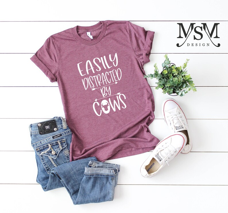 May include: A maroon t-shirt with the text "EASILY DISTRACTED BY COWS" in white. The shirt is paired with blue jeans and white sneakers. A small potted plant is also in the image.