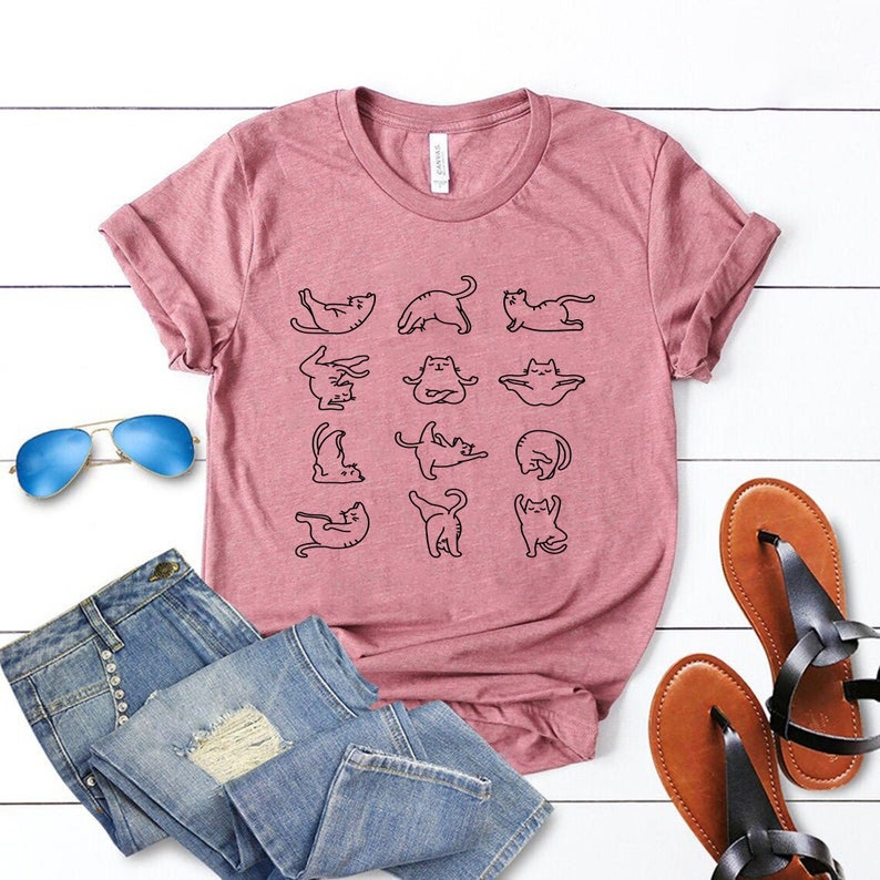 Op de afbeelding: Een roze T-shirt met een zwarte omtrek van katten in verschillende yoga-houdingen. Het shirt ligt op een wit houten oppervlak met een blauwe zonnebril, een blauwe spijkerbroek en bruine sandalen.