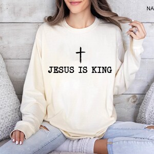K&ouml;nnte beinhalten: Cremefarbenes Langarm-Sweatshirt mit dem schwarzen Text "JESUS IS KING" und einem schwarzen Kreuz dar&uuml;ber. Das Wort "NATURAL" ist in der oberen rechten Ecke aufgedruckt.