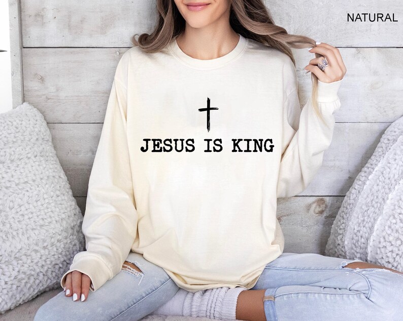 K&ouml;nnte beinhalten: Cremefarbenes Langarm-Sweatshirt mit dem schwarzen Text "JESUS IS KING" und einem schwarzen Kreuz dar&uuml;ber. Das Wort "NATURAL" ist in der oberen rechten Ecke aufgedruckt.