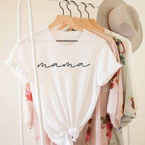 Puede incluir: Una camiseta blanca con la palabra "mama" en escritura negra. La camiseta est&aacute; anudada a la cintura y cuelga de una percha de madera. Otras prendas de vestir y un sombrero tambi&eacute;n est&aacute;n en el perchero.
