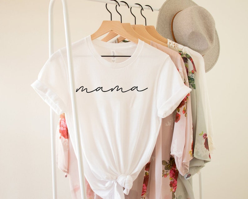 Puede incluir: Una camiseta blanca con la palabra "mama" en escritura negra. La camiseta est&aacute; anudada a la cintura y cuelga de una percha de madera. Otras prendas de vestir y un sombrero tambi&eacute;n est&aacute;n en el perchero.