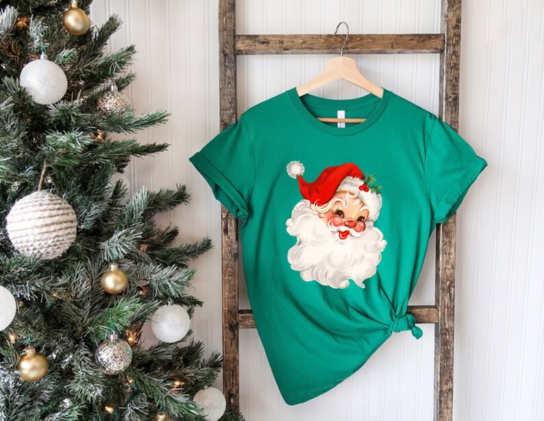 May include: A green t-shirt featuring a vintage Santa Claus graphic. The Santa Claus illustration has a red hat, rosy cheeks, and a white beard. The shirt is hanging on a wooden hanger against a white background.