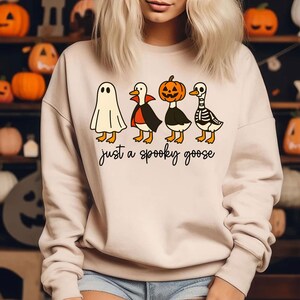 Puede incluir: Sudadera beige con un dise&ntilde;o de Halloween. El dise&ntilde;o muestra cuatro gansos de dibujos animados disfrazados de fantasma, vampiro, calabaza y esqueleto. El texto "just a spooky goose" est&aacute; debajo.
