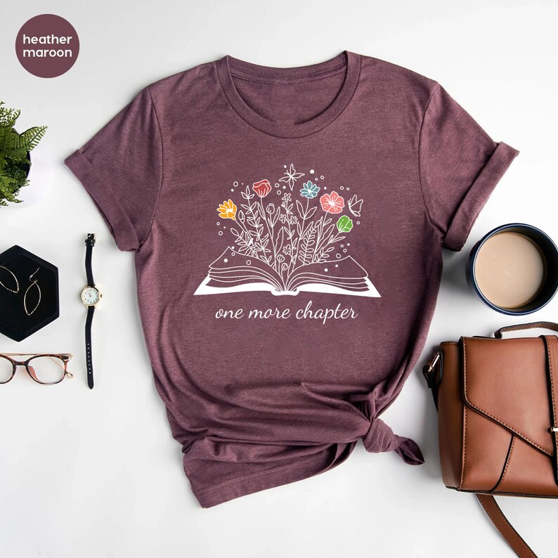 May include: A heather maroon t-shirt with a white graphic of an open book with colorful flowers growing out of it. The text "one more chapter" is printed below the book.