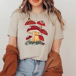 K&ouml;nnte beinhalten: Beiges T-Shirt mit einer verspielten Illustration einer orangefarbenen Katze unter roten Pilzen mit wei&szlig;en Punkten. Ein Schmetterling und kleine Blumen erg&auml;nzen das Design. Dieses l&auml;ssige Shirt ist ein Kleidungsst&uuml;ck im Natur-Look.