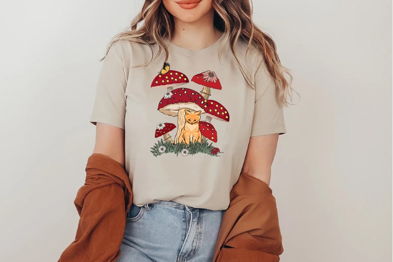 K&ouml;nnte beinhalten: Beiges T-Shirt mit einer verspielten Illustration einer orangefarbenen Katze unter roten Pilzen mit wei&szlig;en Punkten. Ein Schmetterling und kleine Blumen erg&auml;nzen das Design. Dieses l&auml;ssige Shirt ist ein Kleidungsst&uuml;ck im Natur-Look.