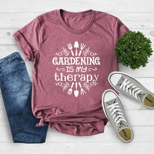 Op de afbeelding: Een maronkleurig T-shirt met witte tekst die "Gardening is my therapy" luidt en tuinwerktuigen en bladeren rond de tekst.