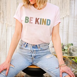 May include: A light pink t-shirt with the words "BE KIND" printed across the front in a multi-colored font. The shirt has rolled-up sleeves and is paired with blue jeans. The overall style is casual and comfortable.