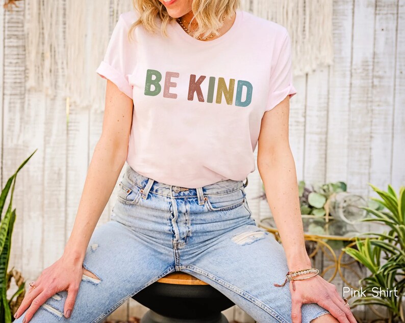 May include: A light pink t-shirt with the words "BE KIND" printed across the front in a multi-colored font. The shirt has rolled-up sleeves and is paired with blue jeans. The overall style is casual and comfortable.