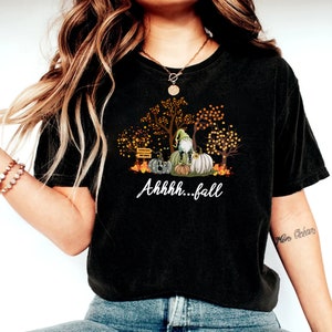 May include: A black t-shirt featuring a graphic print of a gnome sitting on pumpkins surrounded by autumn trees with orange and yellow leaves. The text "Ahhhh...fall" is printed in white script below the graphic, which also includes a small signpost.