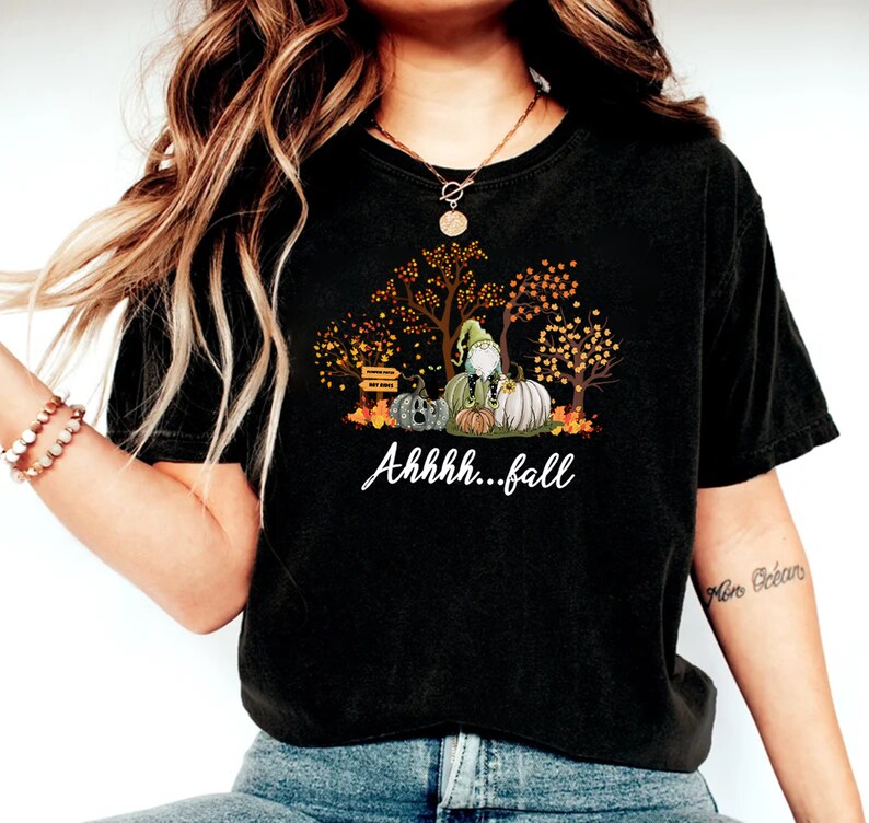 May include: A black t-shirt featuring a graphic print of a gnome sitting on pumpkins surrounded by autumn trees with orange and yellow leaves. The text "Ahhhh...fall" is printed in white script below the graphic, which also includes a small signpost.