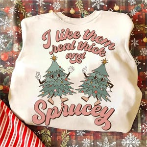 May include: A cream-colored crewneck sweatshirt featuring a graphic of two cartoon Christmas trees with arms and legs. The text reads, I like them real thick and Sprucey, in a retro pink font. The trees are green with colorful ornaments.
