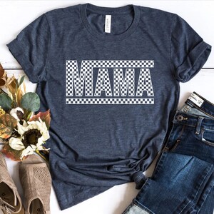 Puede incluir: Camiseta azul oscuro con la palabra "MAMA" en letras blancas a cuadros. La camiseta se combina con unos vaqueros azules y unas sandalias beige. Un ramo de flores de colores oto&ntilde;ales se coloca cerca.