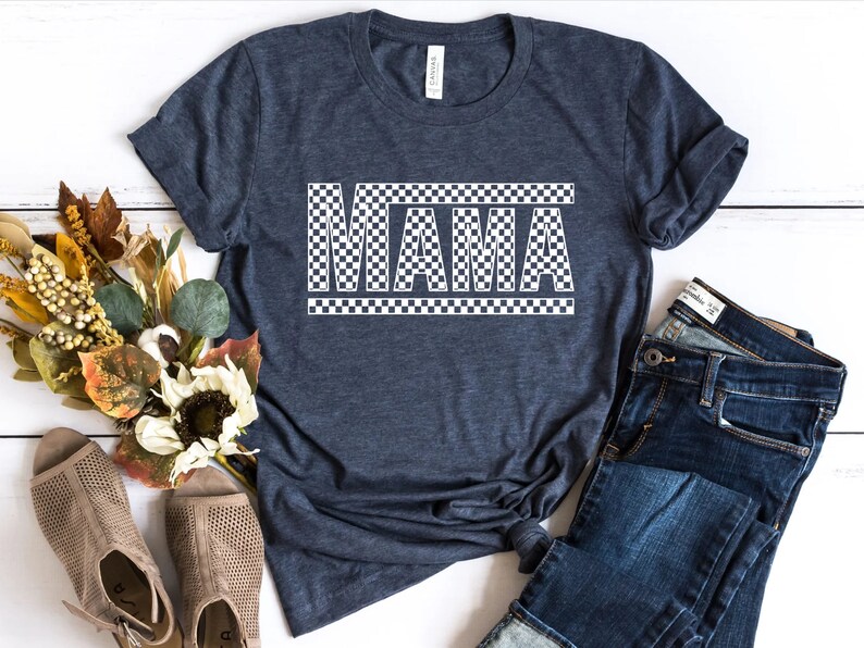 Puede incluir: Camiseta azul oscuro con la palabra "MAMA" en letras blancas a cuadros. La camiseta se combina con unos vaqueros azules y unas sandalias beige. Un ramo de flores de colores oto&ntilde;ales se coloca cerca.