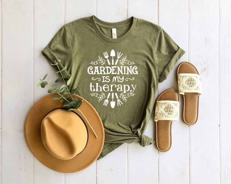Op de afbeelding: Een groen T-shirt met witte tekst die "Tuinieren is mijn therapie" zegt en een afbeelding van tuingereedschap en bladeren.