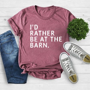 Puede incluir: Una camiseta granate con el texto "I'D RATHER BE AT THE BARN." se muestra con vaqueros azules y zapatillas grises. La camisa est&aacute; doblada en la parte inferior. Una peque&ntilde;a planta verde est&aacute; en el fondo.