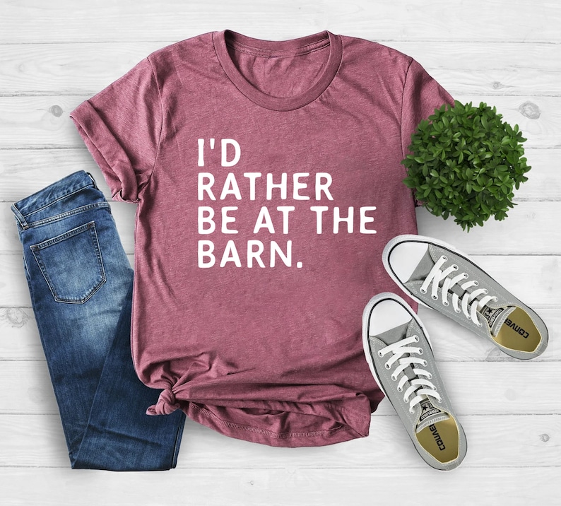 Puede incluir: Una camiseta granate con el texto "I'D RATHER BE AT THE BARN." se muestra con vaqueros azules y zapatillas grises. La camisa est&aacute; doblada en la parte inferior. Una peque&ntilde;a planta verde est&aacute; en el fondo.