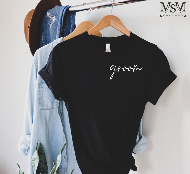 K&ouml;nnte beinhalten: Ein schwarzes T-Shirt mit dem Wort "groom" in wei&szlig;er Schrift. Das Shirt h&auml;ngt an einem Holzkleiderb&uuml;gel, mit einem hellblauen Jeanshemd und einem braunen Hut im Hintergrund. Das Shirt ist Teil eines Hochzeitsthemas.