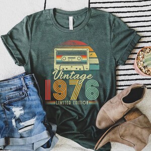 May include: A dark green t-shirt featuring a graphic of a cassette tape above the text Vintage 1976 Limited Edition in a retro, multi-coloured striped font. The design includes a sunset motif behind the cassette tape, rendered in shades of orange, yellow, and teal.
