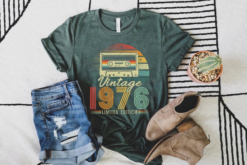 May include: A dark green t-shirt featuring a graphic of a cassette tape above the text Vintage 1976 Limited Edition in a retro, multi-coloured striped font. The design includes a sunset motif behind the cassette tape, rendered in shades of orange, yellow, and teal.