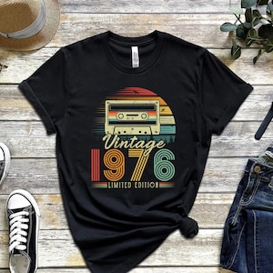 May include: A black short-sleeved t-shirt featuring a graphic of a cassette tape centred above a sunset design. The text reads "Vintage 1976 LIMITED EDITION" in a retro font. The shirt is displayed on a wooden surface with accessories.
