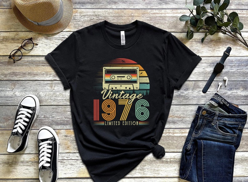 May include: A black short-sleeved t-shirt featuring a graphic of a cassette tape centred above a sunset design. The text reads "Vintage 1976 LIMITED EDITION" in a retro font. The shirt is displayed on a wooden surface with accessories.