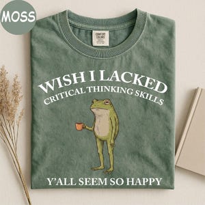 May include: A moss green crew neck t-shirt featuring a graphic of a standing frog holding a small orange mug. The text reads WISH I LACKED CRITICAL THINKING SKILLS above the frog and Y'ALL SEEM SO HAPPY below the frog in white lettering.