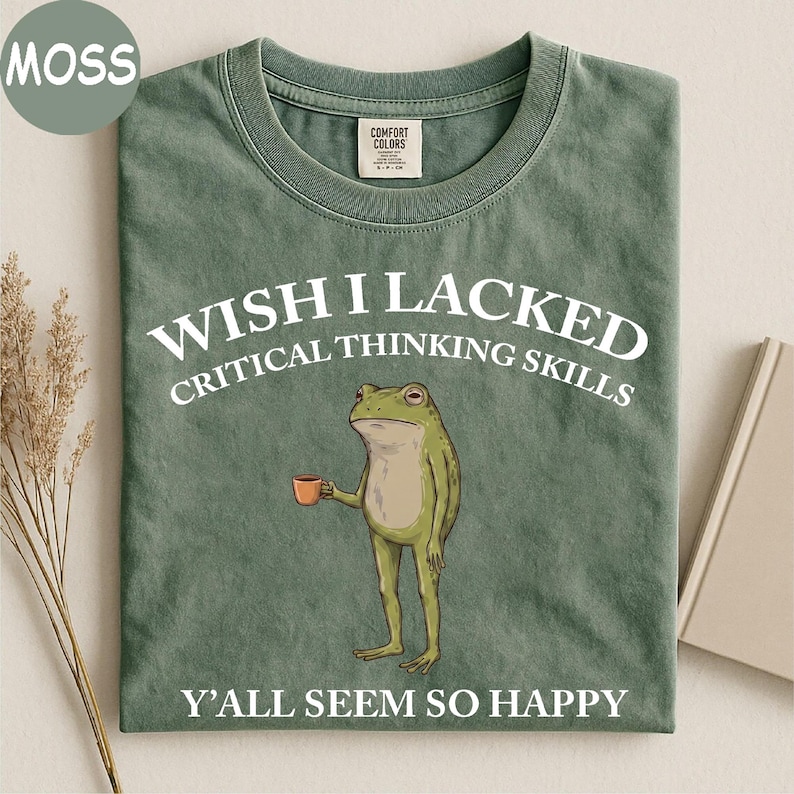 May include: A moss green crew neck t-shirt featuring a graphic of a standing frog holding a small orange mug. The text reads WISH I LACKED CRITICAL THINKING SKILLS above the frog and Y'ALL SEEM SO HAPPY below the frog in white lettering.