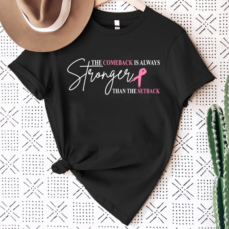 May include: Black t-shirt with the text "THE COMEBACK IS ALWAYS STRONGER THAN THE SETBACK" in white and pink. The design includes a pink ribbon. A tan hat is visible in the upper left corner.