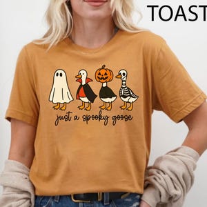 Puede incluir: Camiseta de color mostaza con un dise&ntilde;o de Halloween. El gr&aacute;fico presenta cuatro gansos de dibujos animados disfrazados de fantasma, vampiro, calabaza y esqueleto. El texto "just a spooky goose" est&aacute; debajo.