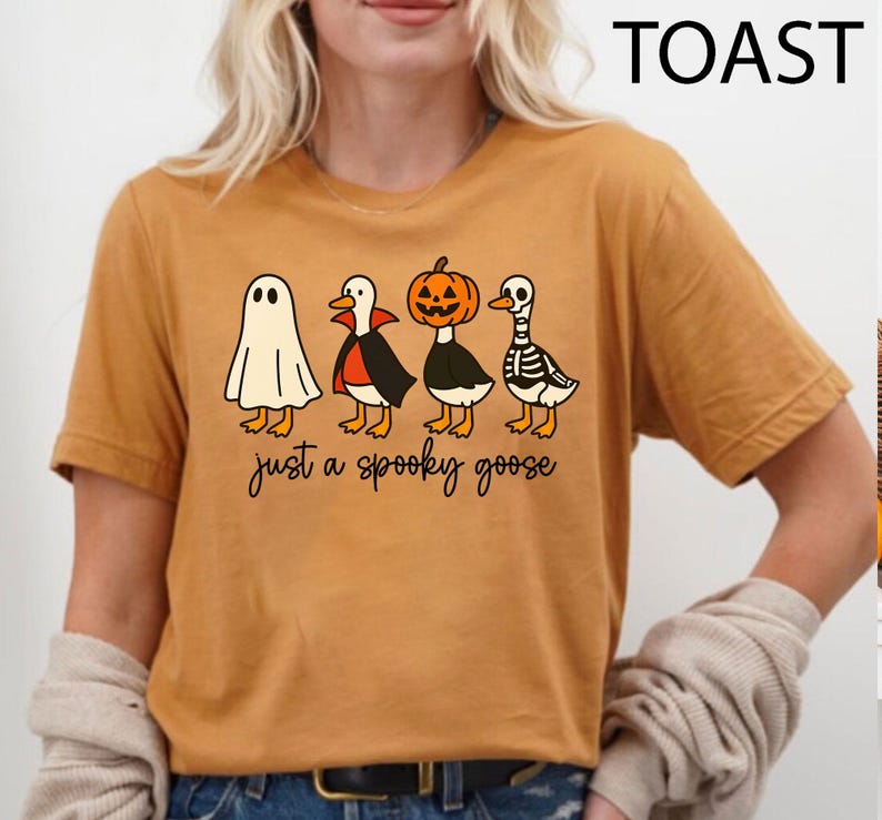 Puede incluir: Camiseta de color mostaza con un dise&ntilde;o de Halloween. El gr&aacute;fico presenta cuatro gansos de dibujos animados disfrazados de fantasma, vampiro, calabaza y esqueleto. El texto "just a spooky goose" est&aacute; debajo.