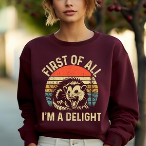 K&ouml;nnte beinhalten: Burgunderfarbenes Sweatshirt mit einem Retro-Grafikdesign eines Opossums in einem Kreis. Der Text lautet "FIRST OF ALL I'M A DELIGHT". Das Design beinhaltet einen Sonnenuntergang.