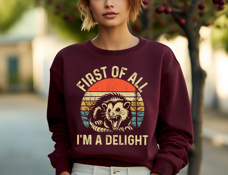 K&ouml;nnte beinhalten: Burgunderfarbenes Sweatshirt mit einem Retro-Grafikdesign eines Opossums in einem Kreis. Der Text lautet "FIRST OF ALL I'M A DELIGHT". Das Design beinhaltet einen Sonnenuntergang.