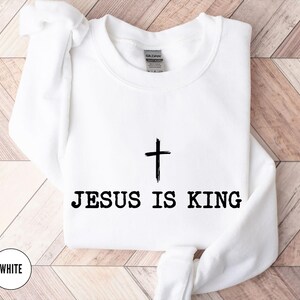 K&ouml;nnte beinhalten: Wei&szlig;es Sweatshirt mit Rundhalsausschnitt und dem schwarzen Text &bdquo;JESUS IS KING&ldquo; unter einer schwarzen Kreuzgrafik. Das Wort &bdquo;WHITE&ldquo; befindet sich in einem wei&szlig;en Kreis unten links. Das Sweatshirt befindet sich auf einer hellbraunen Holzoberfl&auml;che.