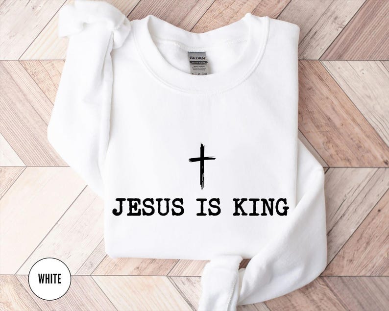 K&ouml;nnte beinhalten: Wei&szlig;es Sweatshirt mit Rundhalsausschnitt und dem schwarzen Text &bdquo;JESUS IS KING&ldquo; unter einer schwarzen Kreuzgrafik. Das Wort &bdquo;WHITE&ldquo; befindet sich in einem wei&szlig;en Kreis unten links. Das Sweatshirt befindet sich auf einer hellbraunen Holzoberfl&auml;che.