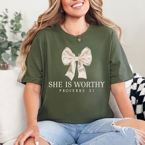 May include: Olive green t-shirt featuring a large, cream-colored bow graphic. The text "SHE IS WORTHY" and "PROVERBS 31" are printed below the bow in a clean, sans-serif font. The shirt is a casual, everyday wear item.