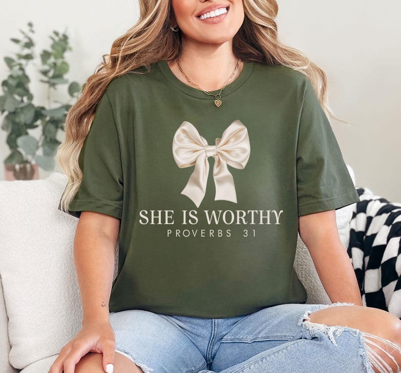 May include: Olive green t-shirt featuring a large, cream-colored bow graphic. The text "SHE IS WORTHY" and "PROVERBS 31" are printed below the bow in a clean, sans-serif font. The shirt is a casual, everyday wear item.
