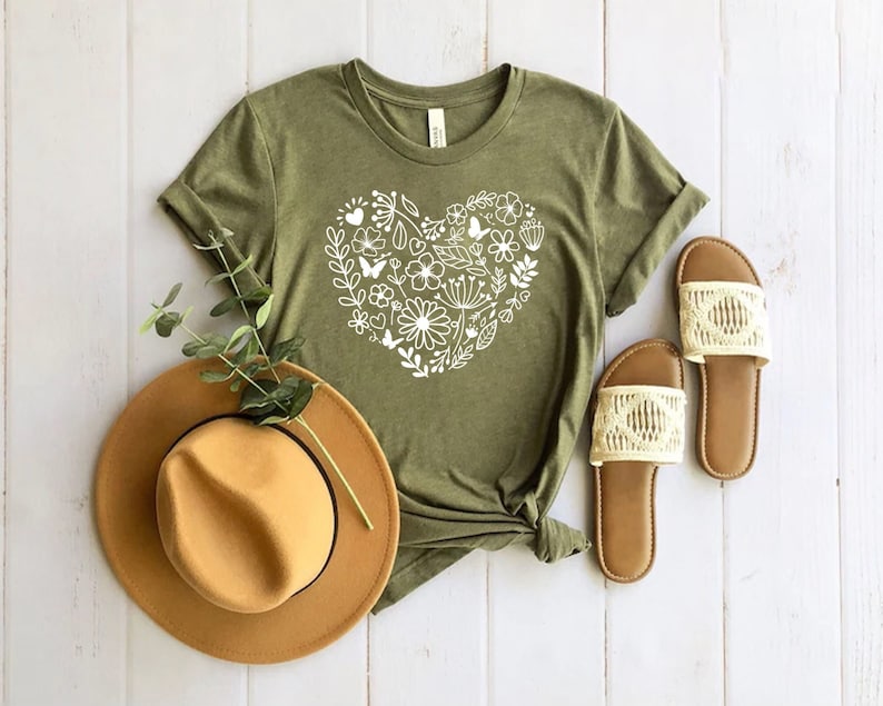 May include: A green t-shirt with a white floral heart design. The shirt is laid out on a white wooden surface with a brown hat and two pairs of sandals.