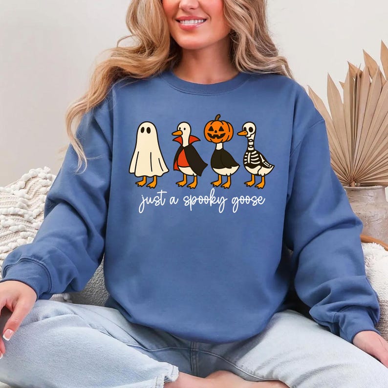 Puede incluir: Sudadera azul con un dise&ntilde;o de Halloween. El gr&aacute;fico muestra cuatro gansos de dibujos animados disfrazados de fantasma, vampiro, calabaza y esqueleto. El texto "just a spooky goose" est&aacute; escrito debajo de los gansos en cursiva.