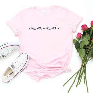 Puede incluir: Una camiseta rosa claro con la palabra "mama" en escritura negra. La imagen tambi&eacute;n presenta zapatillas blancas y un ramo de rosas rosas. La camiseta es de estilo casual.